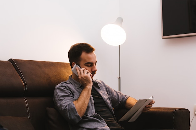 Мужчина держит бумагу и разговаривает по мобильному телефону, сидя на диване у себя дома