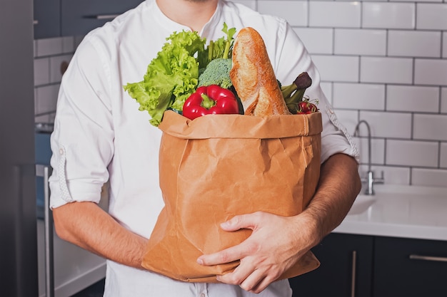 Мужчина держит бумажный пакет с различными овощами и другими продуктами