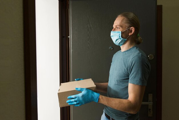 Man holding paper box with food in doors in medical gloves and protective mask copy space safe