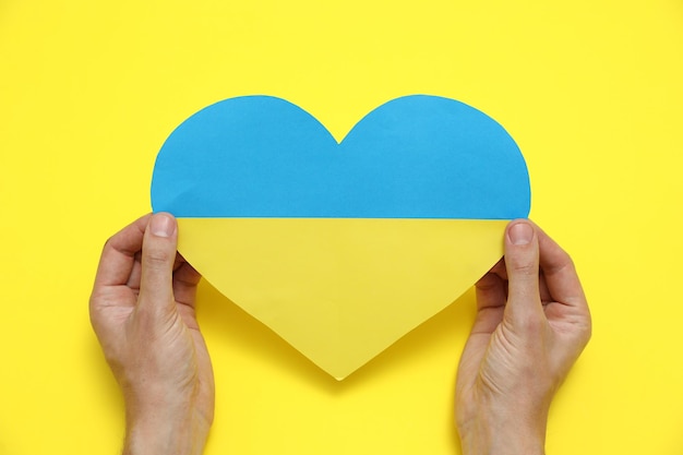 Man holding paper heart in colors of Ukrainian flag on yellow background top view