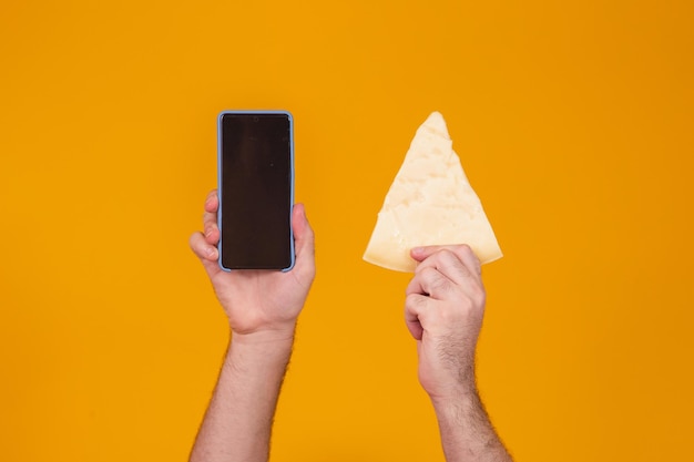 Man holding a piece of cheese in one hand and cellphone in the other with blank screen for text space. Cheese company advertisement concept. Cheese delivery and online shopping