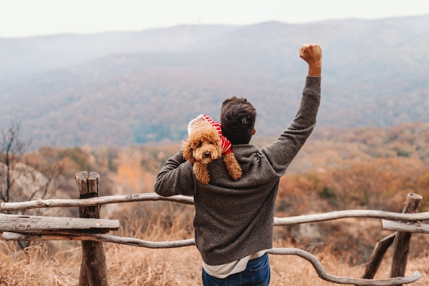 Man holding poodle over the shoulder and lifting fist in the air. Autumn time. Backs turned.