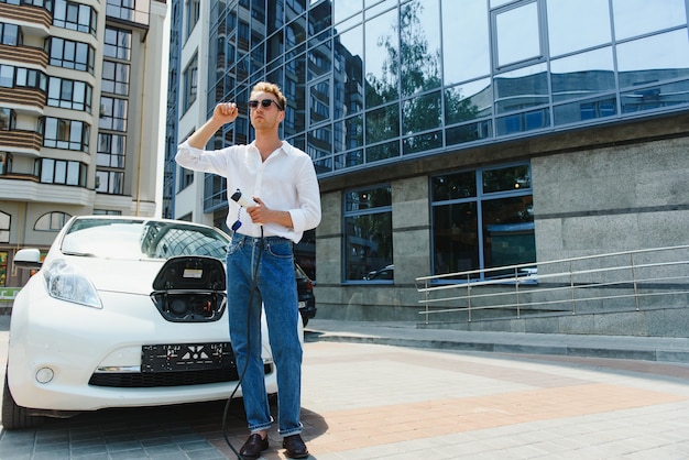 屋外駐車場で電気自動車の充電ケーブルを保持している男性。そして、彼はショッピングセンター近くの駐車場にある充電ステーションに車を接続するつもりです