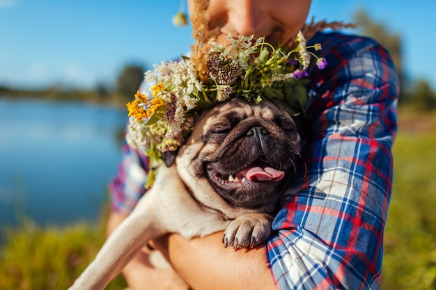 Man holding pug dog with flower wreath on head. Man walking with pet by summer lake