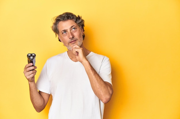 Man holding a razor yellow studio background looking sideways with doubtful and skeptical expression