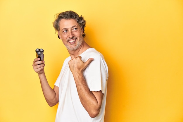 Man holding a razor yellow studio background points with thumb finger away laughing and carefree