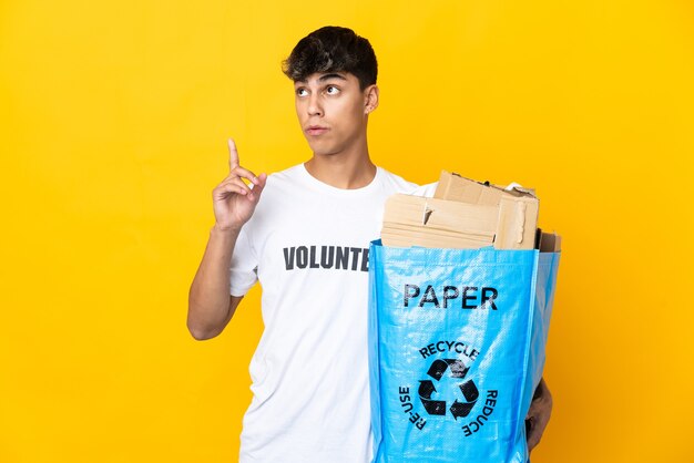 Man holding a recycling bag full of paper to recycle on isolated yellow thinking an idea pointing the finger up