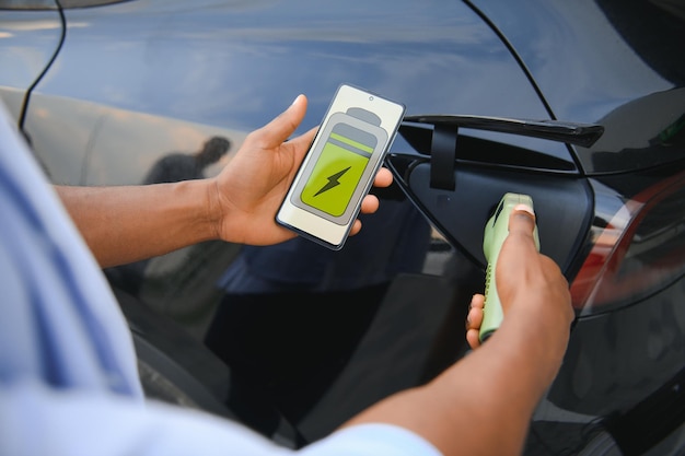 電気自動車の充電ステーションのクローズアップで車を充電しながらスマートフォンを保持している男性