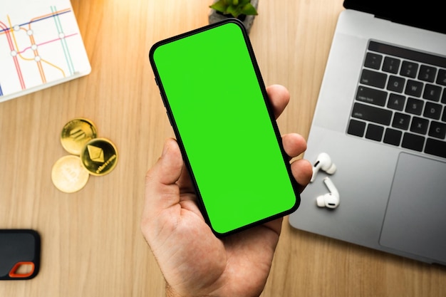 Man holding a smartphone with Green Screen on the screen on wooden table with a cryptocurrencies coins and computer Office environment Chroma Key