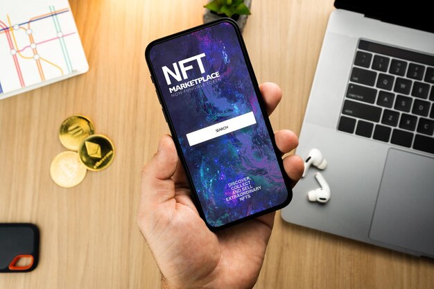 Photo man holding a smartphone with nft nonfungible token marketplace on the screen on wooden table with a cryptocurrencies coins and computer office environment