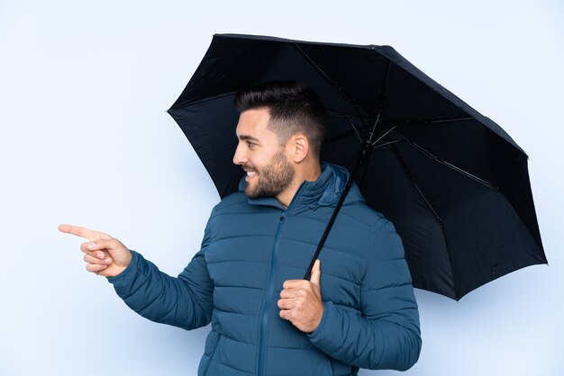 Man holding an umbrella over isolated wall pointing to the side to present a product