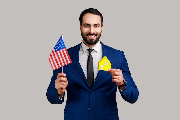 Man holding USA flag and paper house dreaming to buy accommodation in America looking at camera