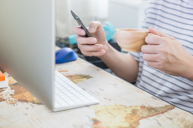 Man holding and using mobile smartphone and holding a wooden mug cup of hot coffee with desktop computer with worldmap mat.