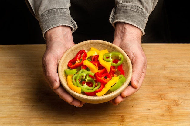 Man holding a wooden bowl with slices of red, green and yellow peppers in a top view