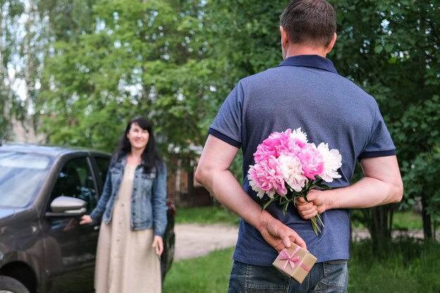 A man holds a bouquet of flowers and a gift behind his back for his girlfriend