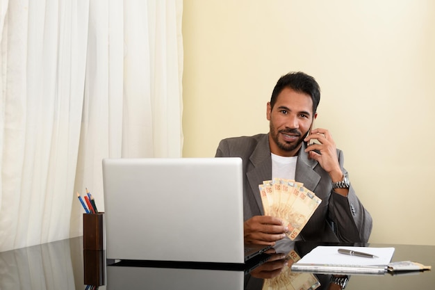 man holds Brazilian money and talks on cell phone, portable computer on table