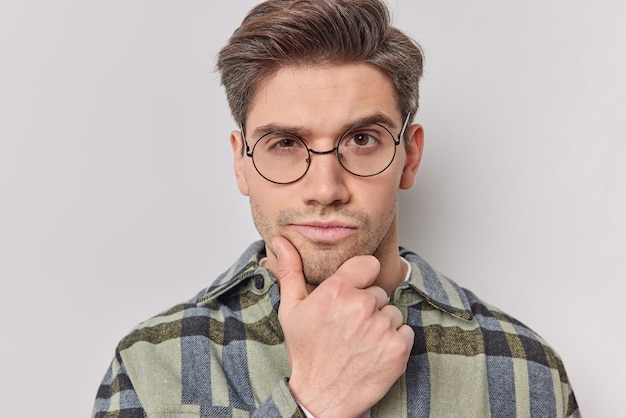 man holds chin looks attentively at camera considers something wears round spectacles and checkered shirt poses on white. Thoughtful handsome guy