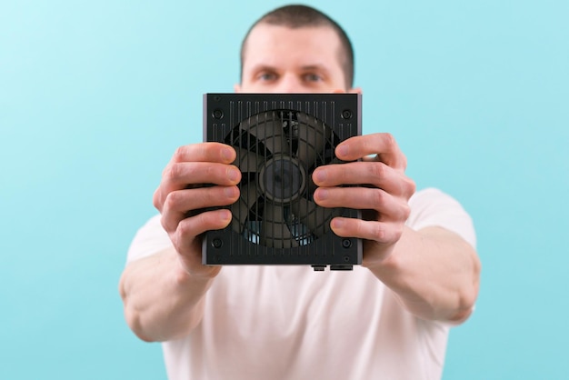 A man holds a computer power supply in front of his face on a\
blue background electrical cooler fan box device part desktop\
peripheral powerful object panel psu energy pc
