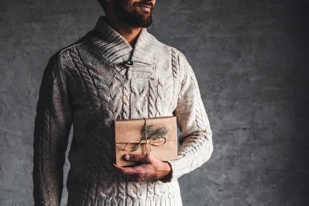 A man holds a craft box for a Christmas present in a beige sweater on a gray background