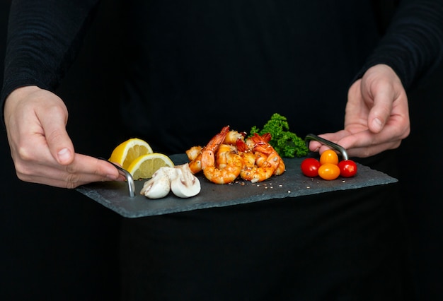 Photo man holds fresh, tasty shrimp on a stone black tray