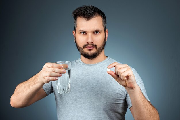 男はコップ1杯の水と錠剤を持ち、薬、治療、灰色の壁を取っています。医療テーマ、ビタミン、ヘルスケア。