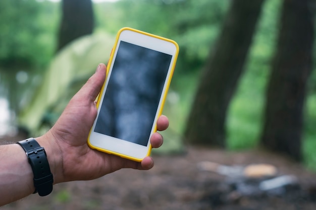 A man holds in his hand a mock-up of a smartphone against the background of nature and a tent.