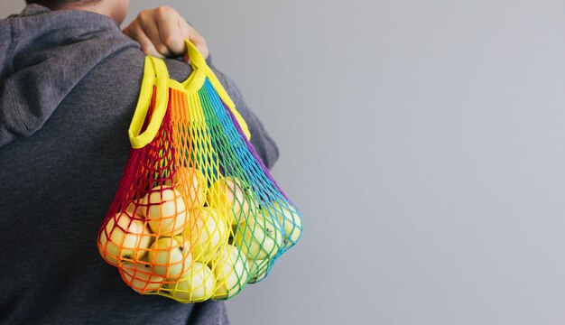 A man holds in his hands a reusable cotton shopping string bag, a multicolored rainbow with green apples inside. gray background. Zero waste concept