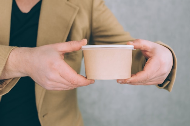 Foto un uomo tiene in mano un modello di un bicchiere di carta per zuppa, caffè e tè su una luce.