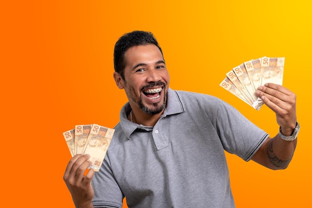 Man holds money, brazilian money in both hands, happy smile, on orange background