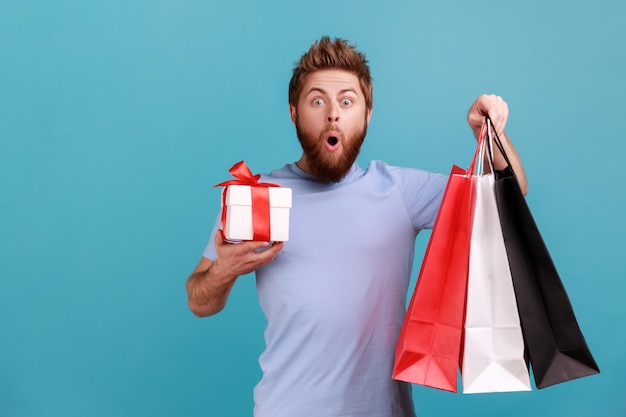 Man holds packages and gift box looks at camera with shocking expression and open mouth saying wow