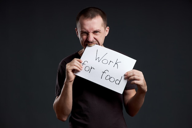 Мужчина держит в руках лист бумаги с надписью «Я работаю за еду». Улыбка и радость, место для текста, копия пространства