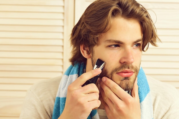 Man holds razor and starts shaving his beard Bearded guy with striped blue towel on light door texture background Barbershop masculine style and morning concept Macho with beard and attentive face