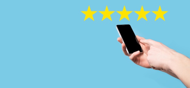 L'uomo tiene lo smart phone nelle mani e dà una valutazione positiva, icona simbolo a cinque stelle per aumentare la valutazione del concetto di azienda su sfondo blu. esperienza di servizio clienti e sondaggio sulla soddisfazione aziendale.