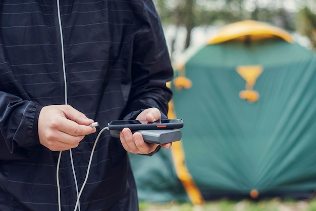 L'uomo tiene in mano uno smartphone e lo carica con una banca di alimentazione sullo sfondo di una tenda turistica nella natura caricabatterie portatile da viaggio