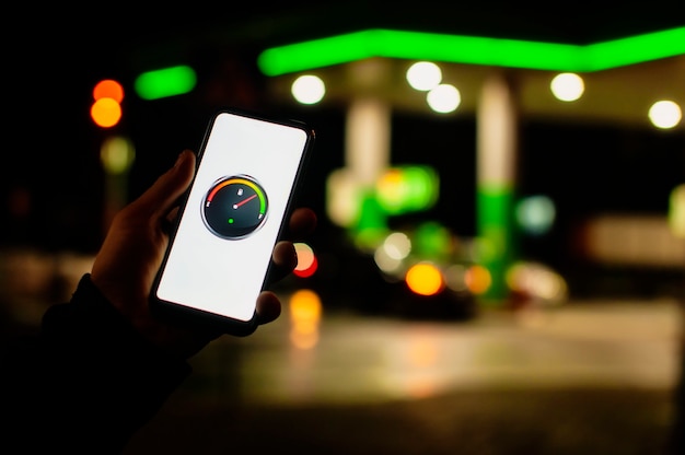 man holds a smartphone with a digital fuel meter on the screen against the background of a night gas station for a car.