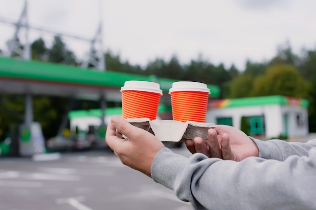 男はガソリンスタンドで彼の手に2杯のコーヒーを持っています