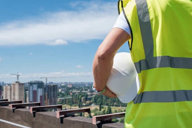 Man holds white hard hat in his hand on the roof of building under construction