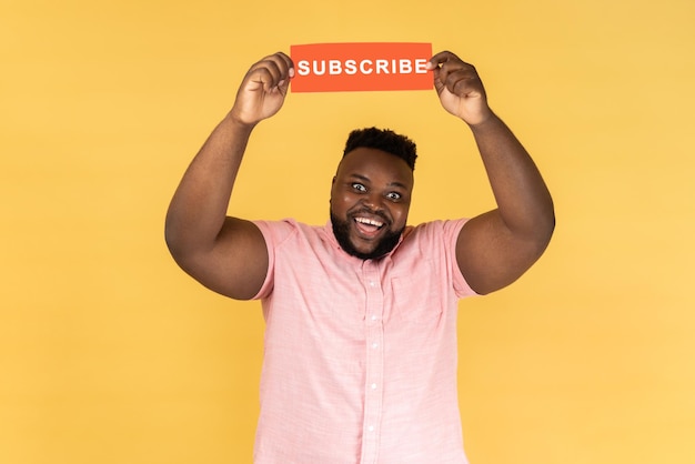 Man holds with paper card with subscribe inscription above head looking at camera with toothy smile