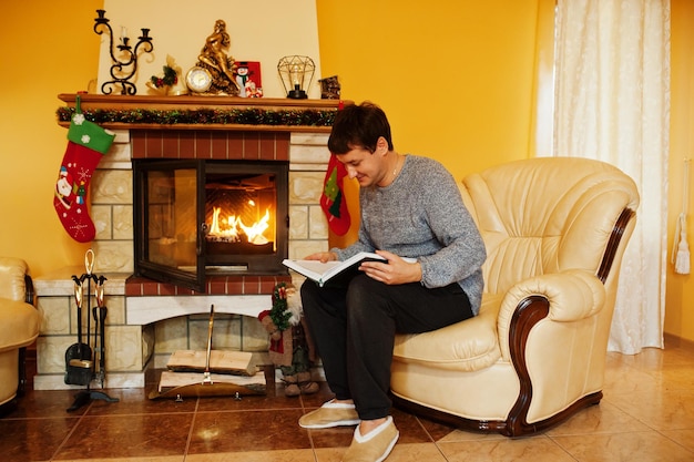 Photo man at home reading book by a fireplace in warm living room on winter day.