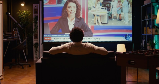 Man in home theatre watching huge TV screen showing news broadcast reportages on family trips