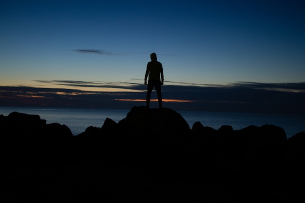 夜の時間に海を背景に岩の上に立っているフードの男