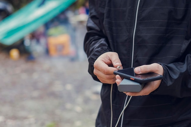 Man houdt een smartphone in zijn handen en laadt deze op met een powerbank tegen de achtergrond van een toeristentent in de natuur Draagbare reislader