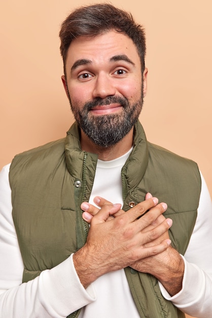 Foto man houdt handen aan het hart maakt dankbaar gebaar waardeert goede houding ten opzichte van hem aangeraakt en blij te zijn gekleed in trui en vest poseert op beige