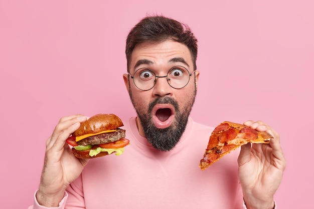 man houdt stuk pizza vast en hamburger eet junkfood geschokt om veel calorieën te consumeren draagt ronde bril casual trui