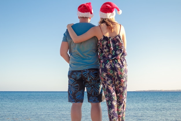 Photo man hugging woman in christmas hats and looking at the beach of the sea.