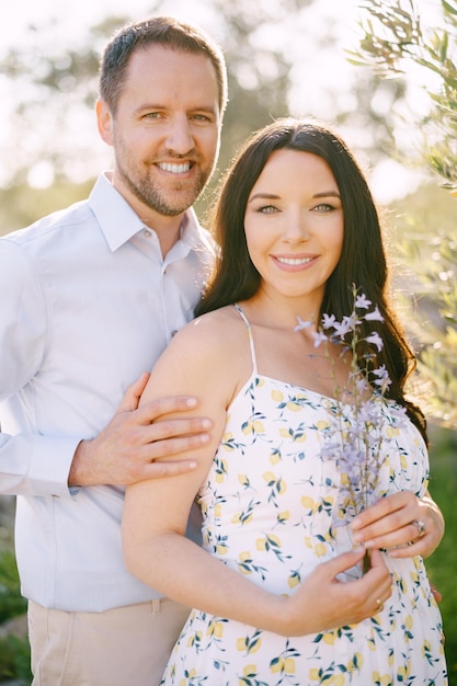 Man hugs pregnant woman holding wildflowers by the shoulders