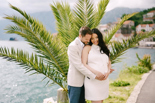Мужчина обнимает улыбающуюся беременную женщину у моря у пальмы