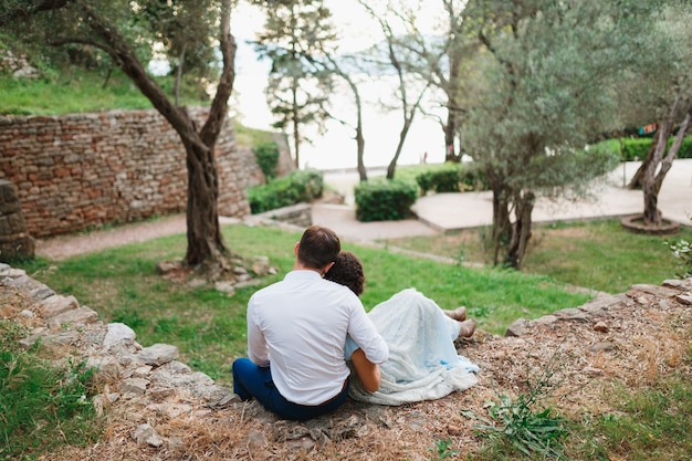 Man hugs woman by the shoulders while sitting on a stone fence between green trees in an olive grove