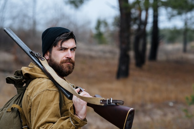 Un uomo cacciatore con barba e baffi in un cappello nero e un grande zaino da trekking tiene una pistola sulla spalla nella foresta