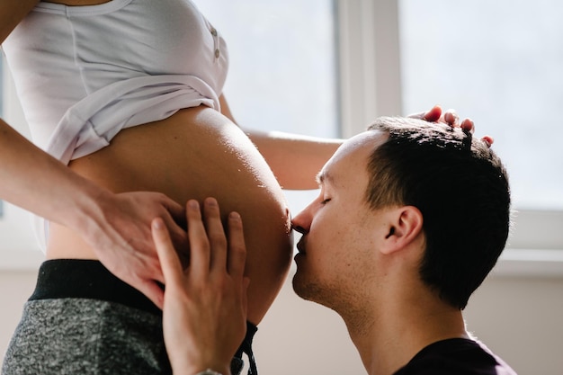 남자 남편은 집 창문 근처에서 임신한 아내의 배에 키스한다 행복한 가족 임신 기대
